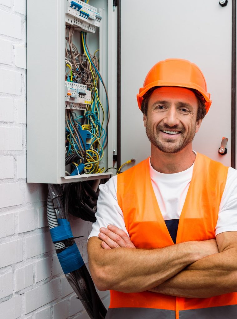 Handsome electrician with crossed arms smiling at camera near electric panel
