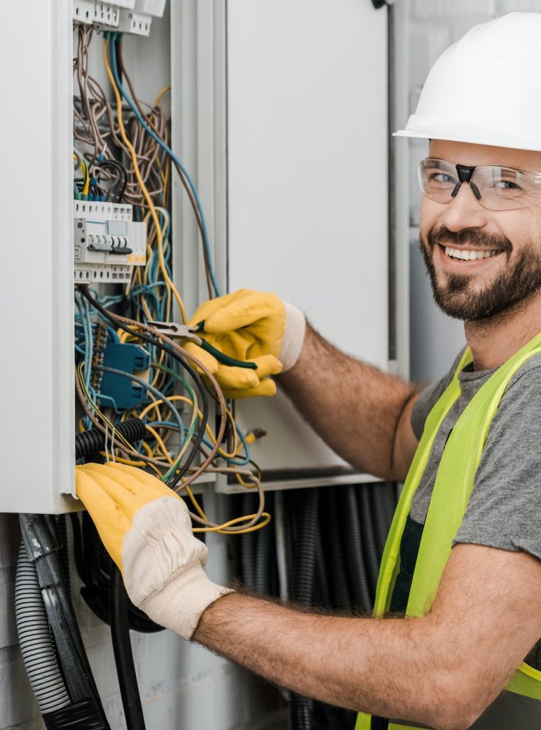 smiling handsome electrician repairing electrical box with pliers in corridor and looking at camera